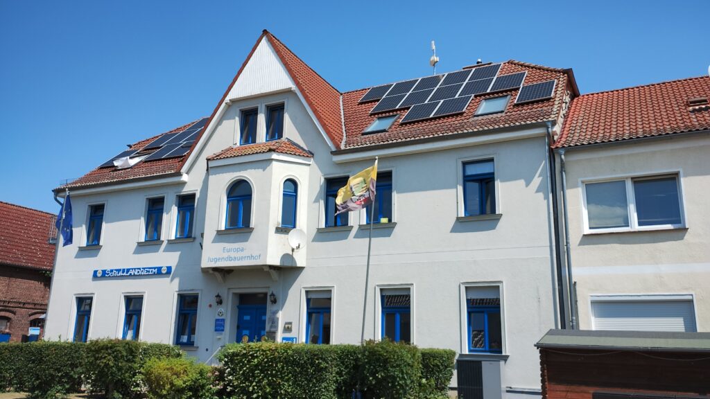 Fassade des Haupteingangs eines Schullandheims und Jugendbauernhofs mit Solarzellen auf rotem Ziegeldach und kleinem Heckengarten bei sonnigem Wetter