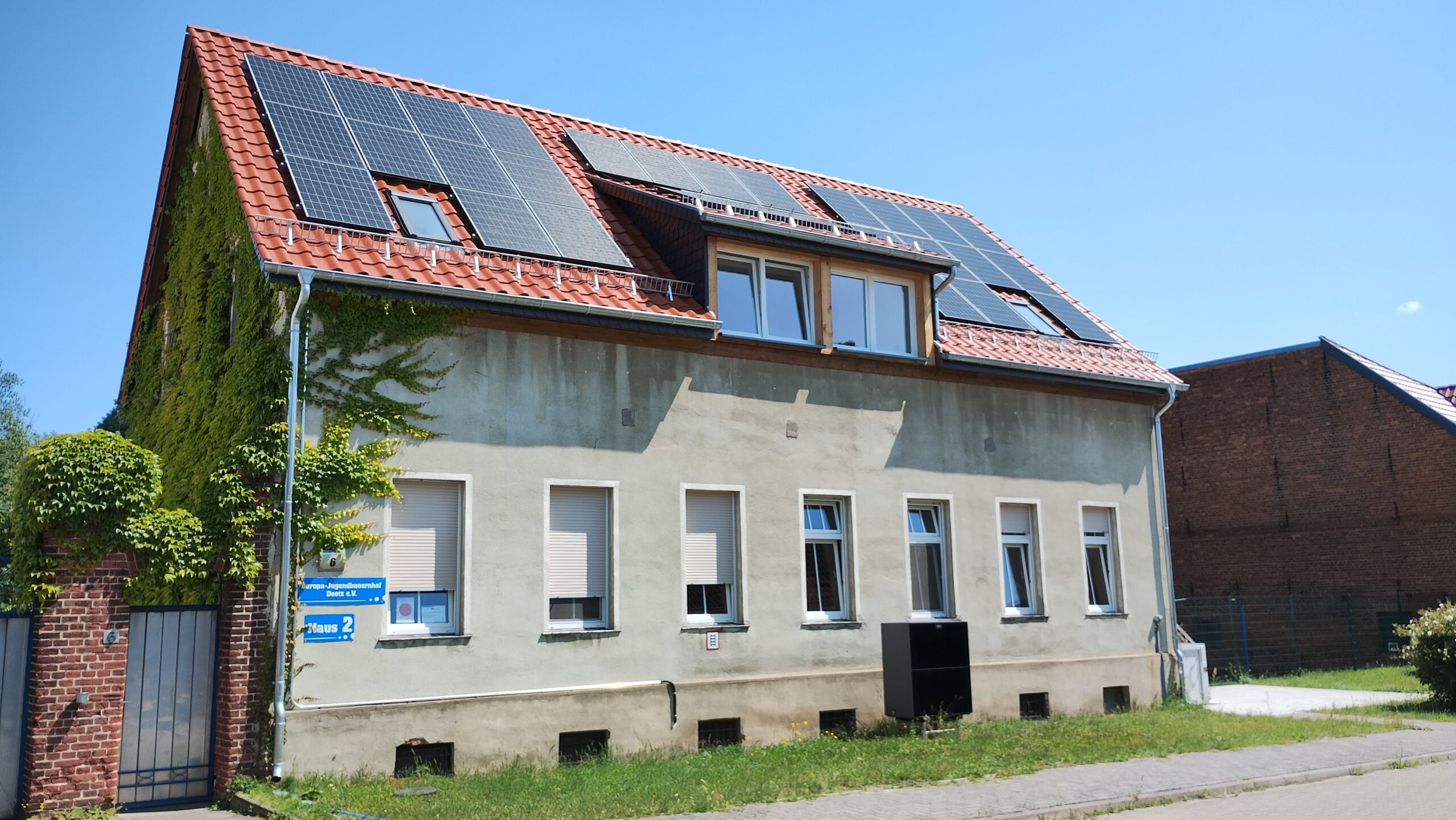 Frontalaufnahme eines Schullandheims und Jugendbauernhofs mit Solarzellen auf rotem Ziegeldach und mit grünem Efeu bewachsener Fassade bei klarem Himmel